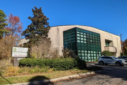 Exterior of modern curved brick treatment facility building with green windows, entrance signage, landscaping, and parking area.