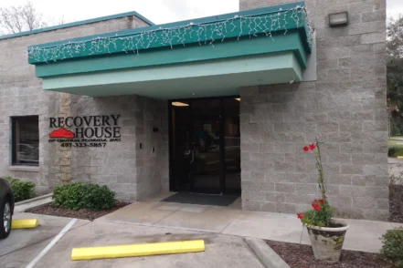 Exterior view of Recovery House of Central Florida showing main entrance with teal awning, stone facade, landscaping, and facility signage with phone number