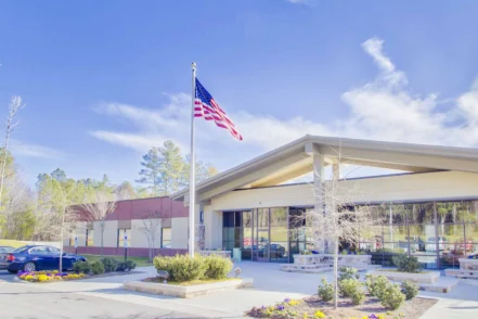 Exterior of modern treatment facility with white and maroon facade, American flag, glass entrance, landscaping, and parking area.
