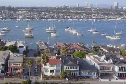 Aerial view of Newport Beach waterfront with sailboats moored in bay, residential homes, palm trees, and neighborhood architecture near water