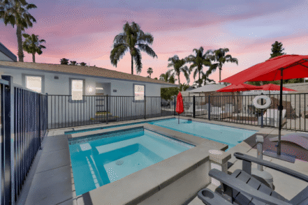 Exterior pool area featuring lap pool, hot tub, lounging chairs, red umbrellas, and facility building with landscaping at dusk